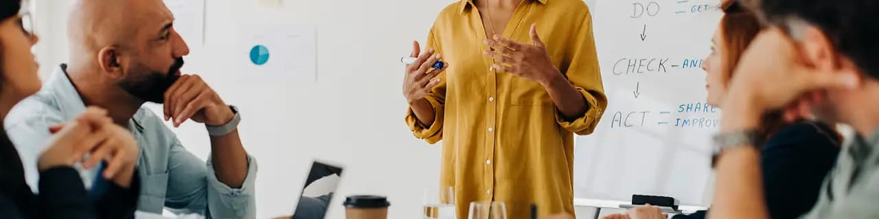 Una donna presenta davanti a una lavagna con le parole DO, CHECK, ACT; diverse persone ascoltano.