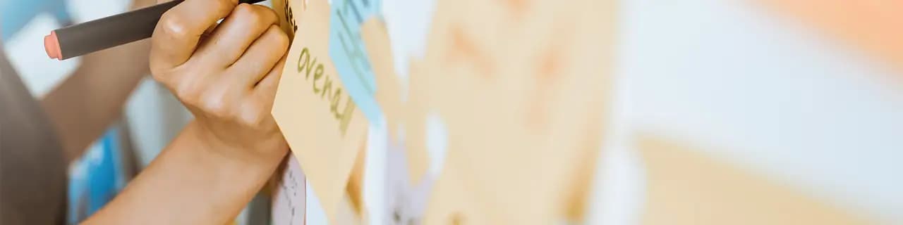 Una mano scrive su un foglietto adesivo su una parete coperta di carte colorate.